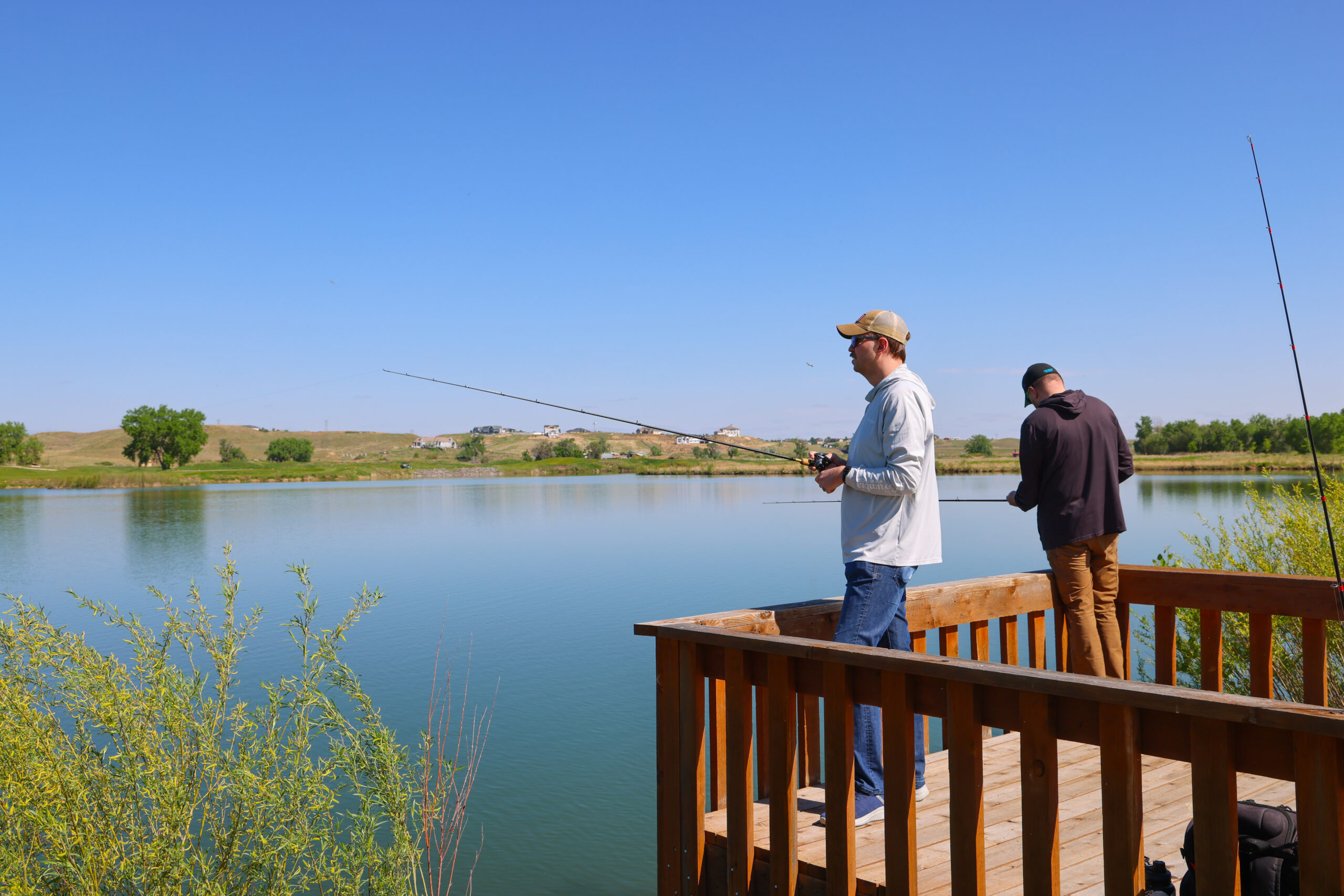 Two people fishing from a wooden platform by a serene lake.