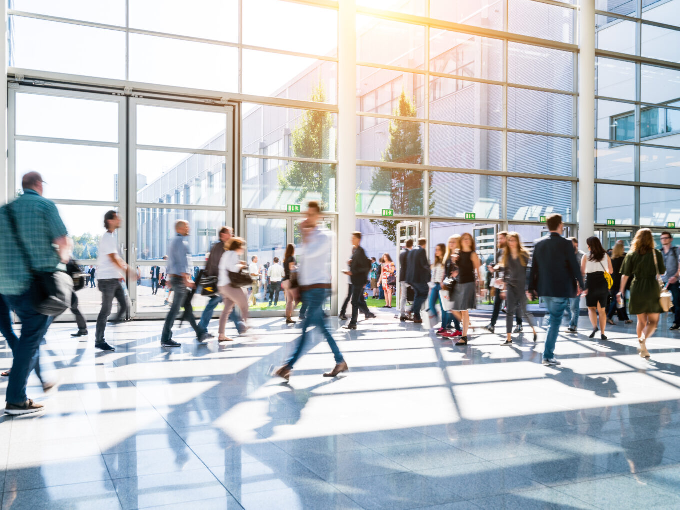 Crowded modern atrium with people walking, large windows, bright sunlight streaming in.
