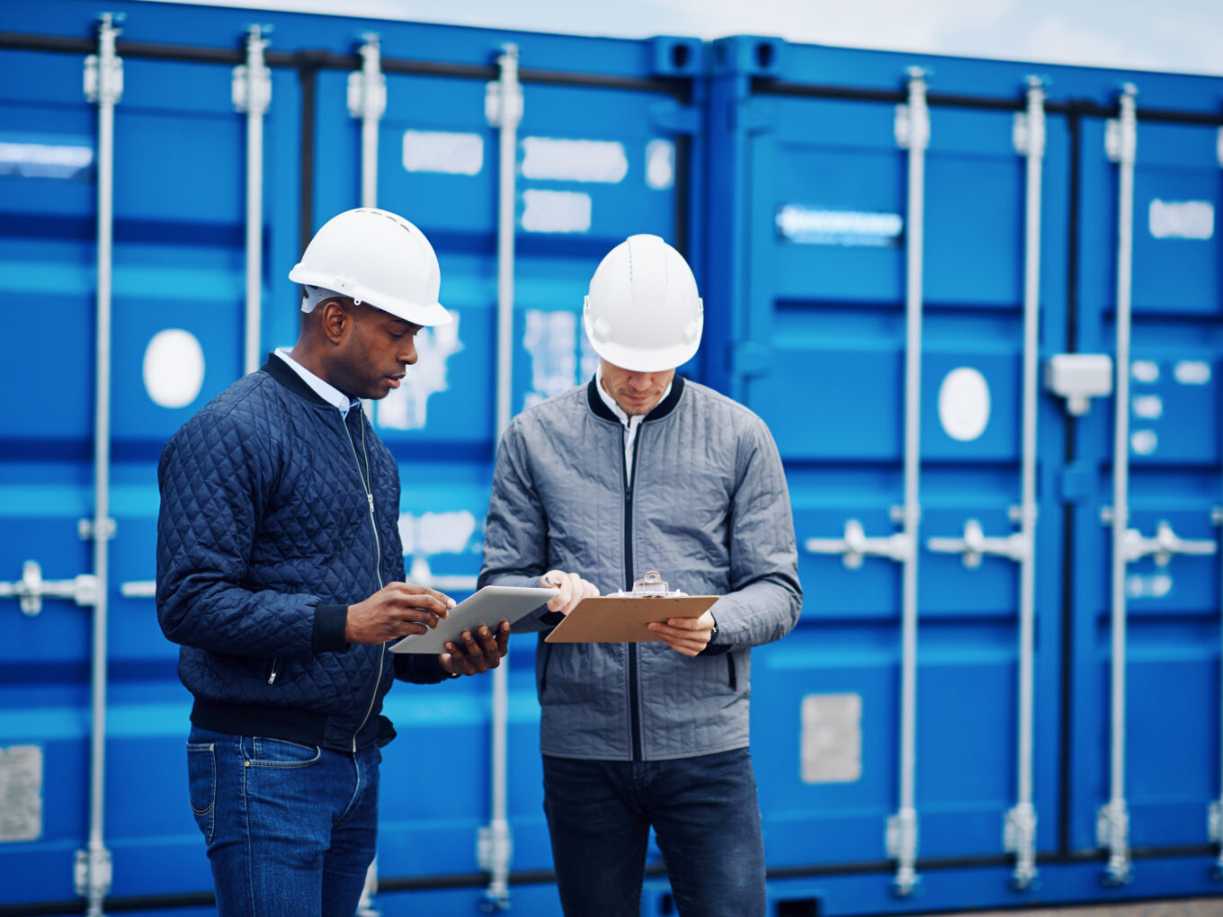 Two workers in hard hats reviewing documents by blue shipping containers.