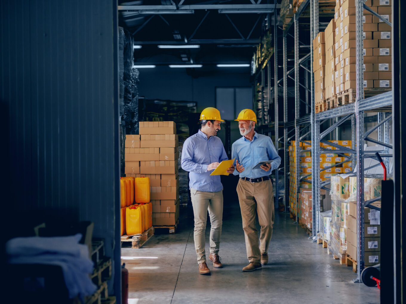 Two men in yellow hard hats walking through a warehouse discussing documents.