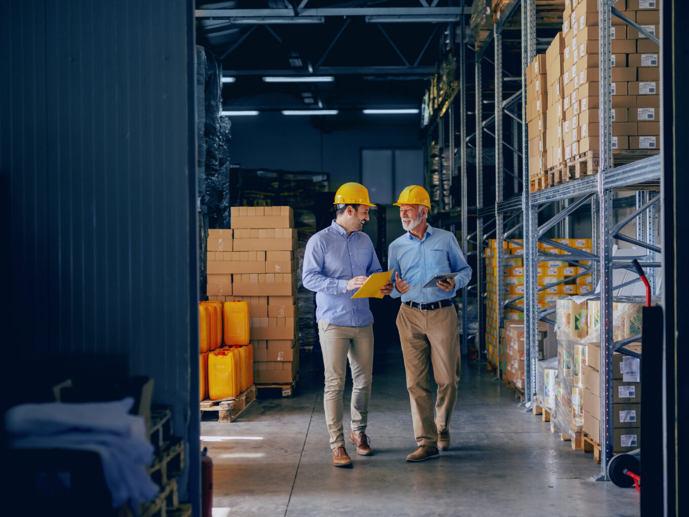 Two men wearing yellow hard hats walking in a warehouse, discussing documents.