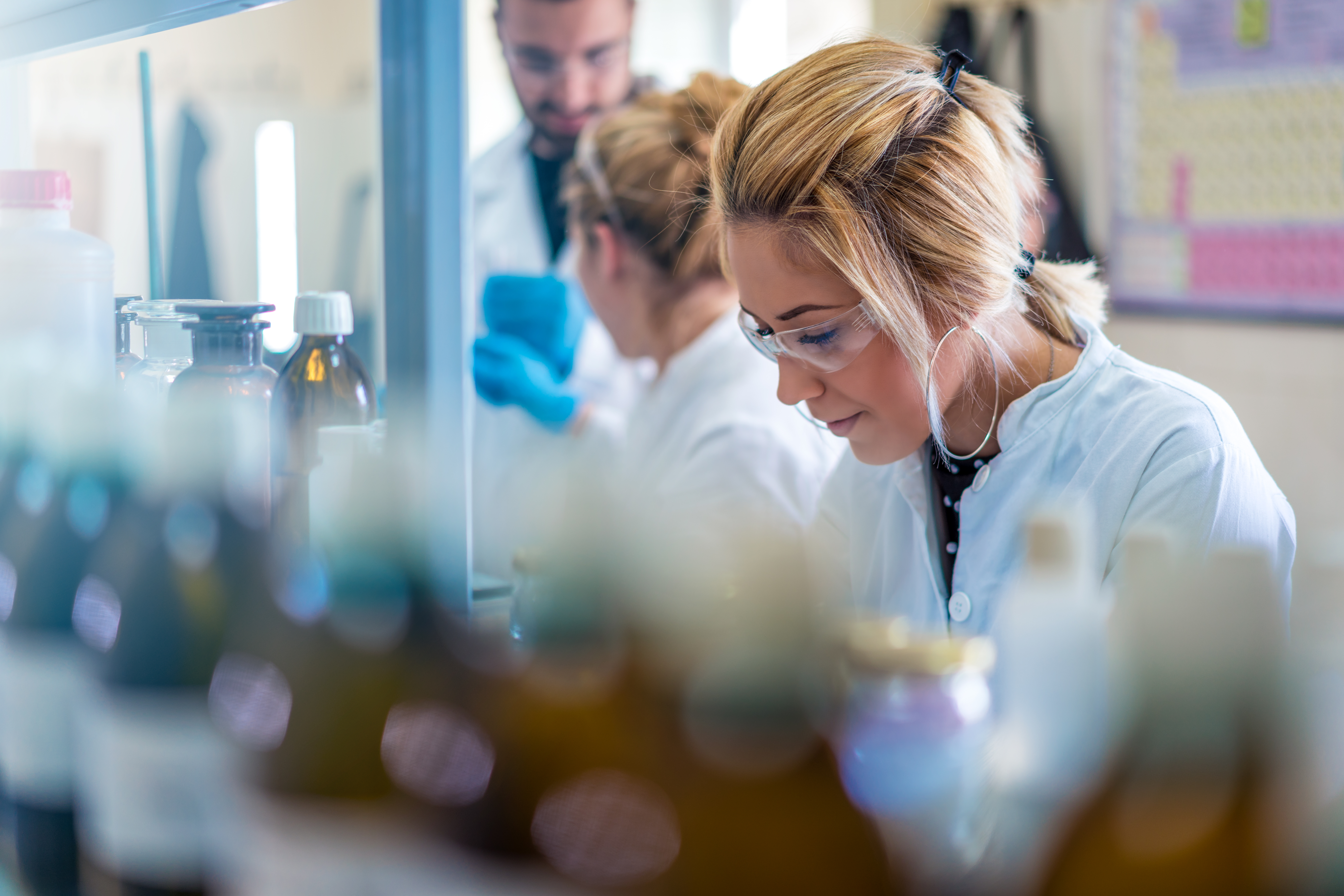 Woman in lab coat and goggles focuses intently, surrounded by bottles.