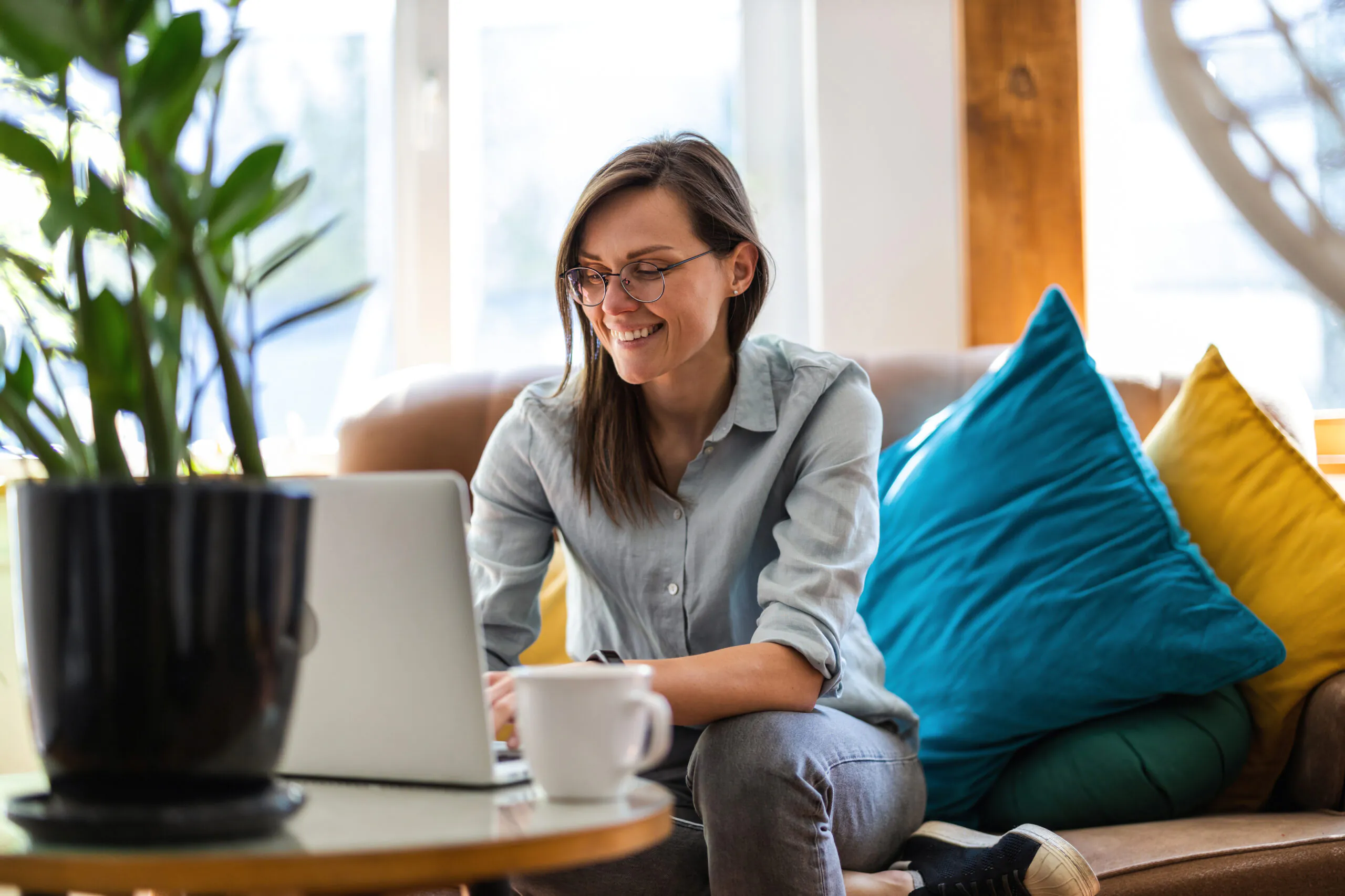 A person smiling while using a laptop on a couch, with colorful pillows and a plant nearby.