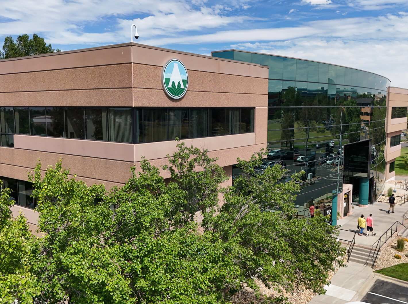 Modern office building with reflective glass facade and mountain logo, surrounded by trees and people walking nearby