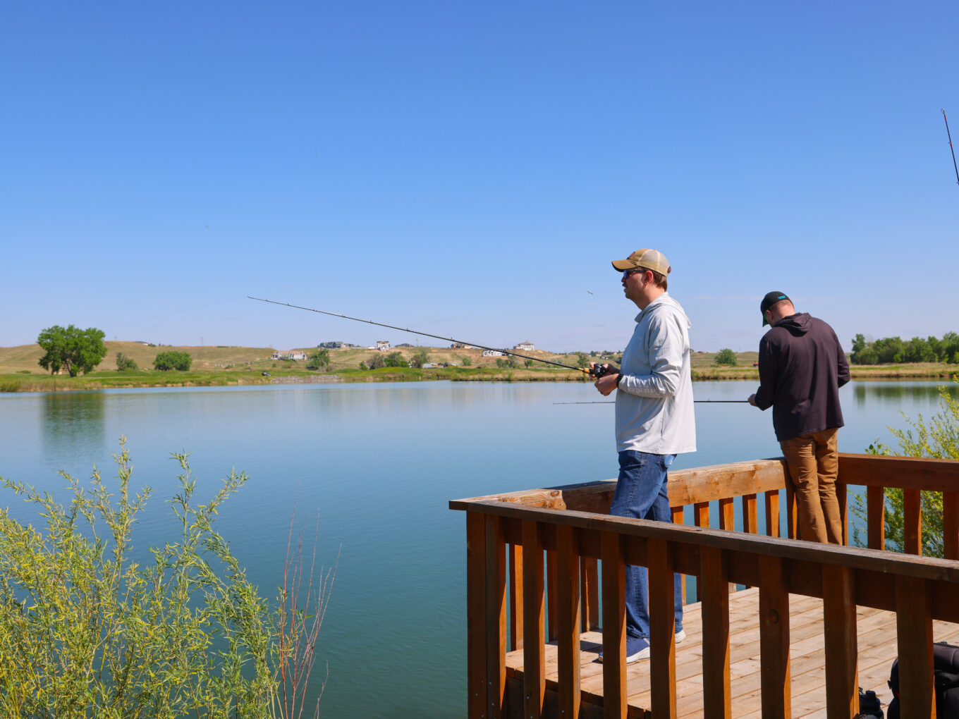 Two people fishing from a wooden platform by a serene lake.