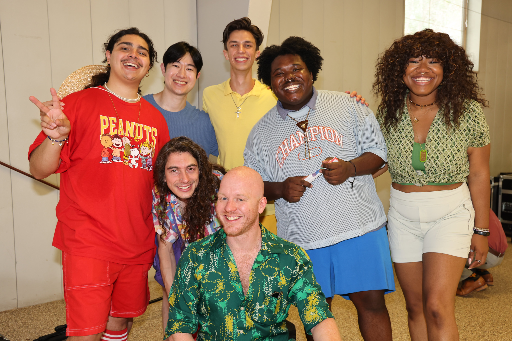 Seven people smiling, one wearing a red "Peanuts" shirt, another a "Champion" shirt, indoors.